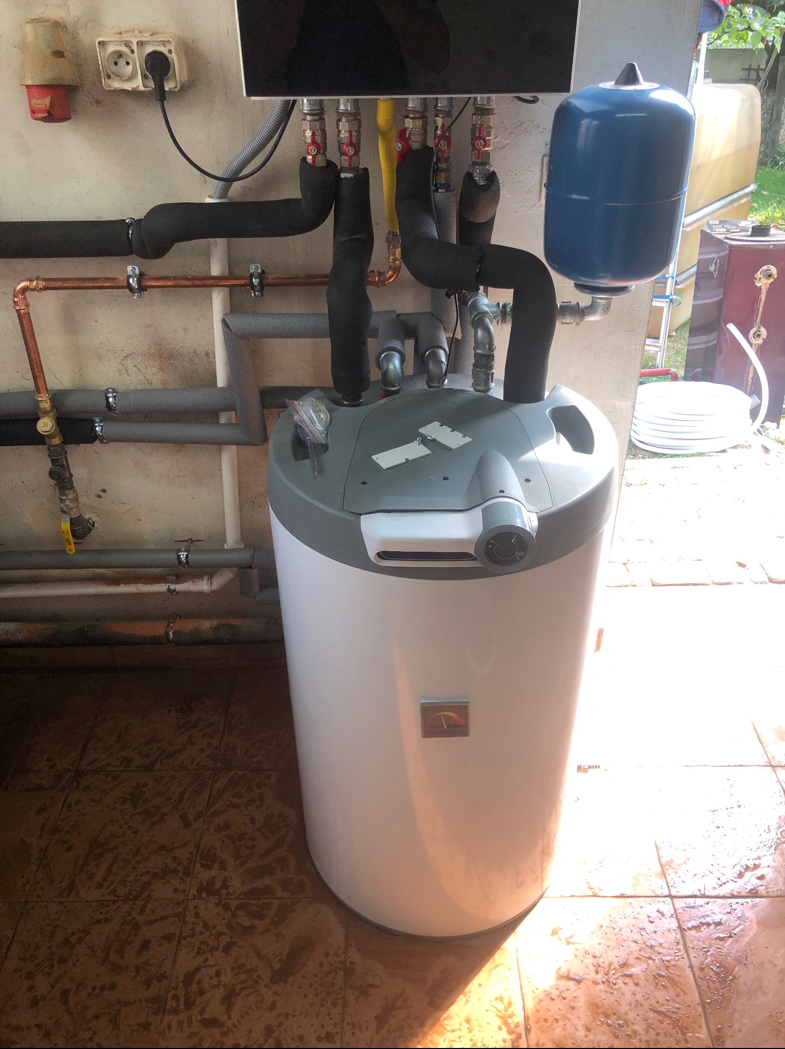 Bojler elektryczny z podłączonymi rurami miedzianymi i izolowanymi, zbiornik wyrównawczy na ścianie, widoczne przyłącza elektryczne i gazowe, na podłodze płytki ceramiczne.