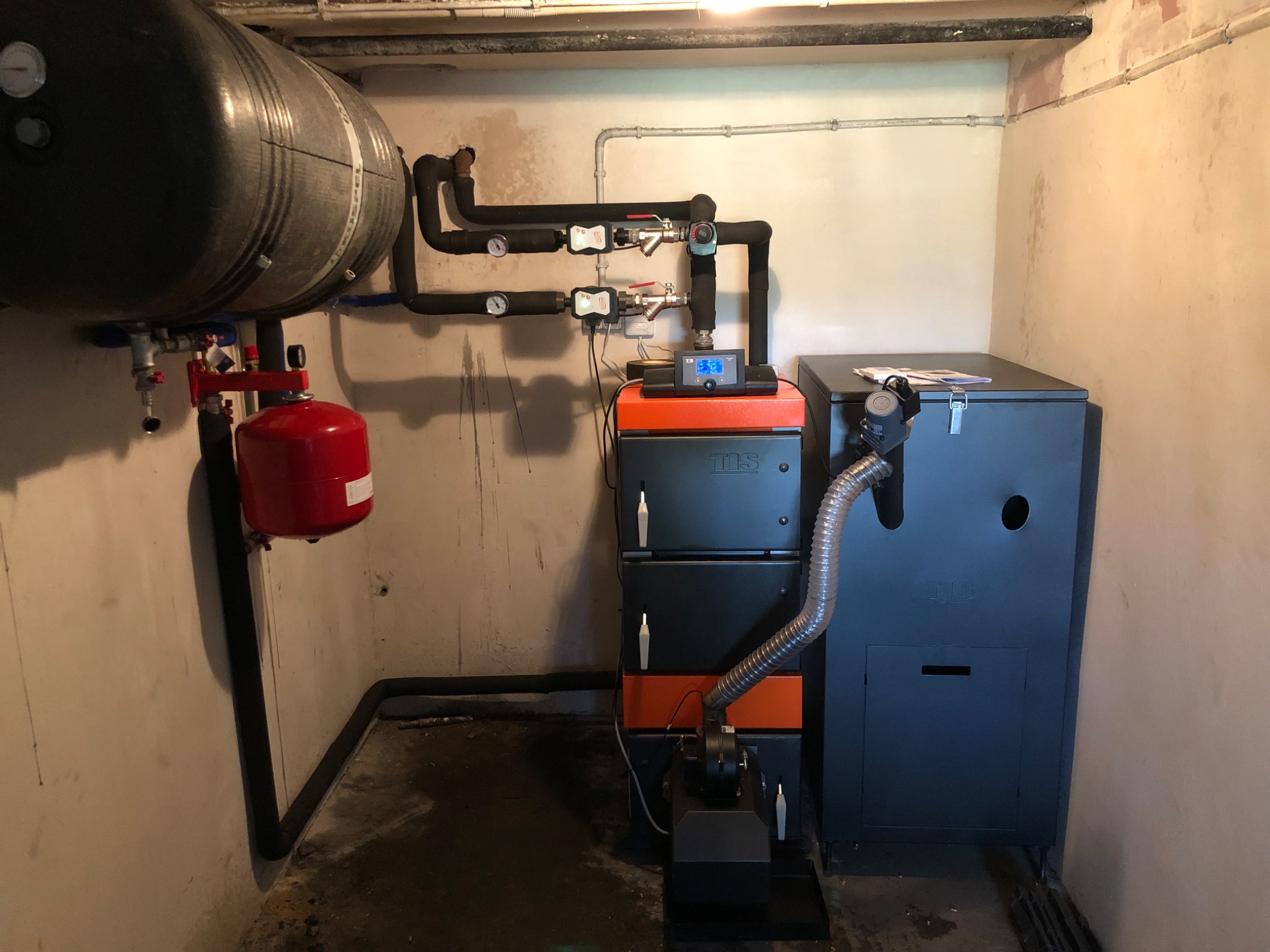 Kotłownia z kotłem na paliwo stałe, zbiornikiem akumulacyjnym i instalacją hydrauliczną w piwnicy, widoczne rury i zawory.