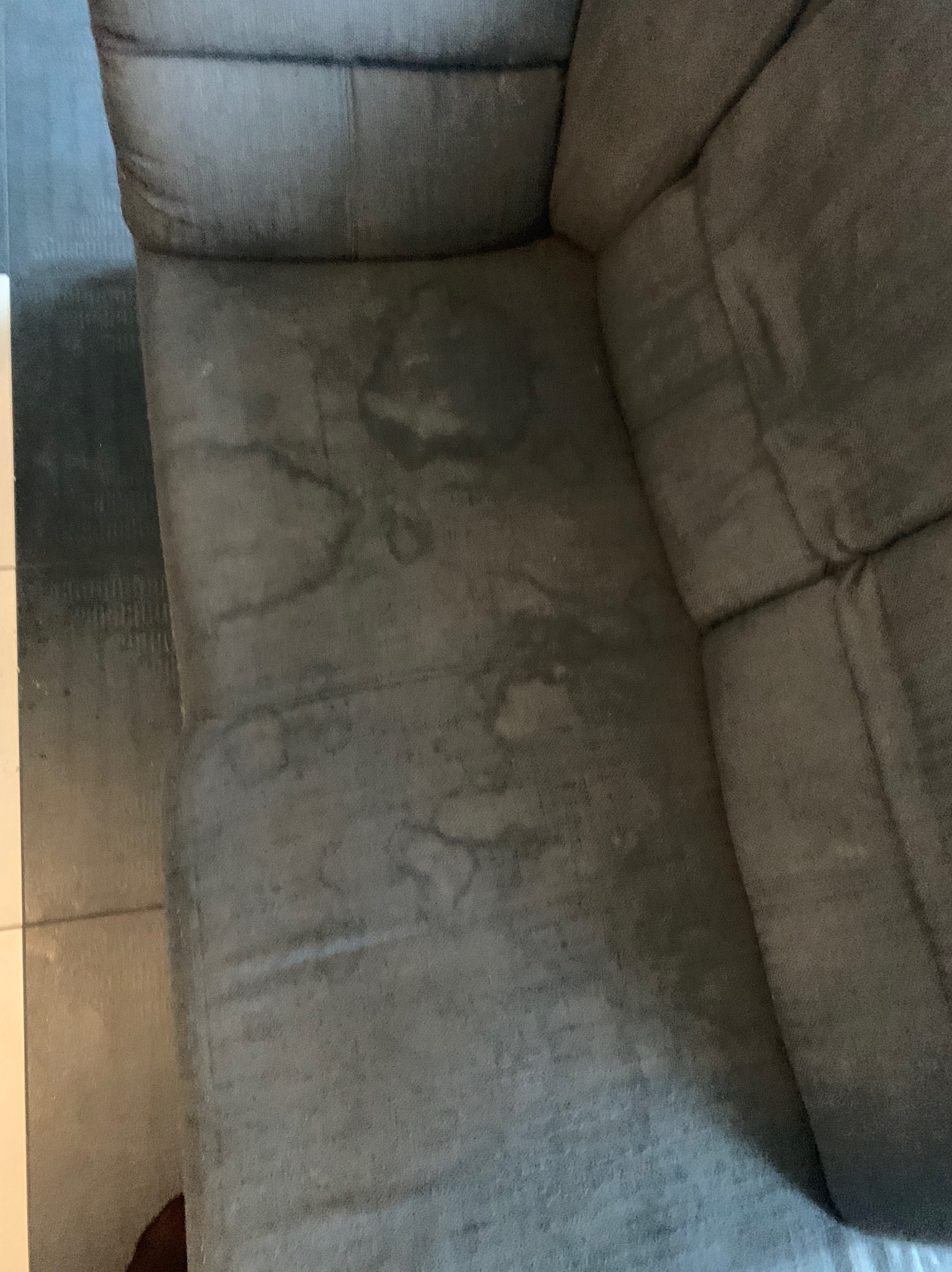 Szara, narożna kanapa tapicerowana z widocznymi, ciemniejszymi plamami i zabrudzeniami na siedzisku i oparciu, sfotografowana z góry przed czyszczeniem.