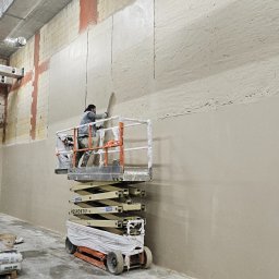 PHU PERFECT BUD MATEUSZ SPYTEK - Pracownik na podnośniku nożycowym wygładza świeżo nałożony tynk na ścianie w surowym wnętrzu budynku, widoczne worki z zaprawą tynkarską.