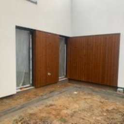 PHU PERFECT BUD MATEUSZ SPYTEK - Nowoczesna elewacja budynku z brązowymi panelami drewnopodobnymi, częściowo zasłaniającymi okno, widoczne świeże ślady prac ziemnych.