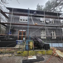 PHU PERFECT BUD MATEUSZ SPYTEK - Dwupiętrowy dom w trakcie prac ociepleniowych elewacji, pokryty czarnym styropianem, z widocznymi rusztowaniami i pracownikami na rusztowaniach, podwórko z gruzem i narzędziami.