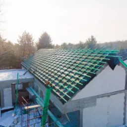 Budowa domu w zimowej scenerii: widok na nieotynkowane ściany z betonu komórkowego i konstrukcję dachu z zielonymi łatami, przygotowaną pod pokrycie.