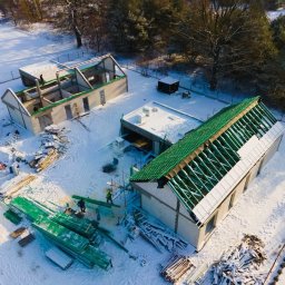 PHU PERFECT BUD MATEUSZ SPYTEK - Zimowy widok z drona na plac budowy z dwoma budynkami w stanie surowym: jeden z konstrukcją dachu w trakcie montażu zielonego pokrycia, drugi w trakcie wznoszenia ścian. Śnieg pokrywa teren...