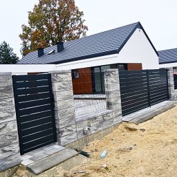 PHU PERFECT BUD MATEUSZ SPYTEK - Nowoczesne ogrodzenie posesji z szarych, betonowych bloczków imitujących kamień i czarnych, poziomych paneli, widok z zewnątrz na tle domu z ciemną dachówką.