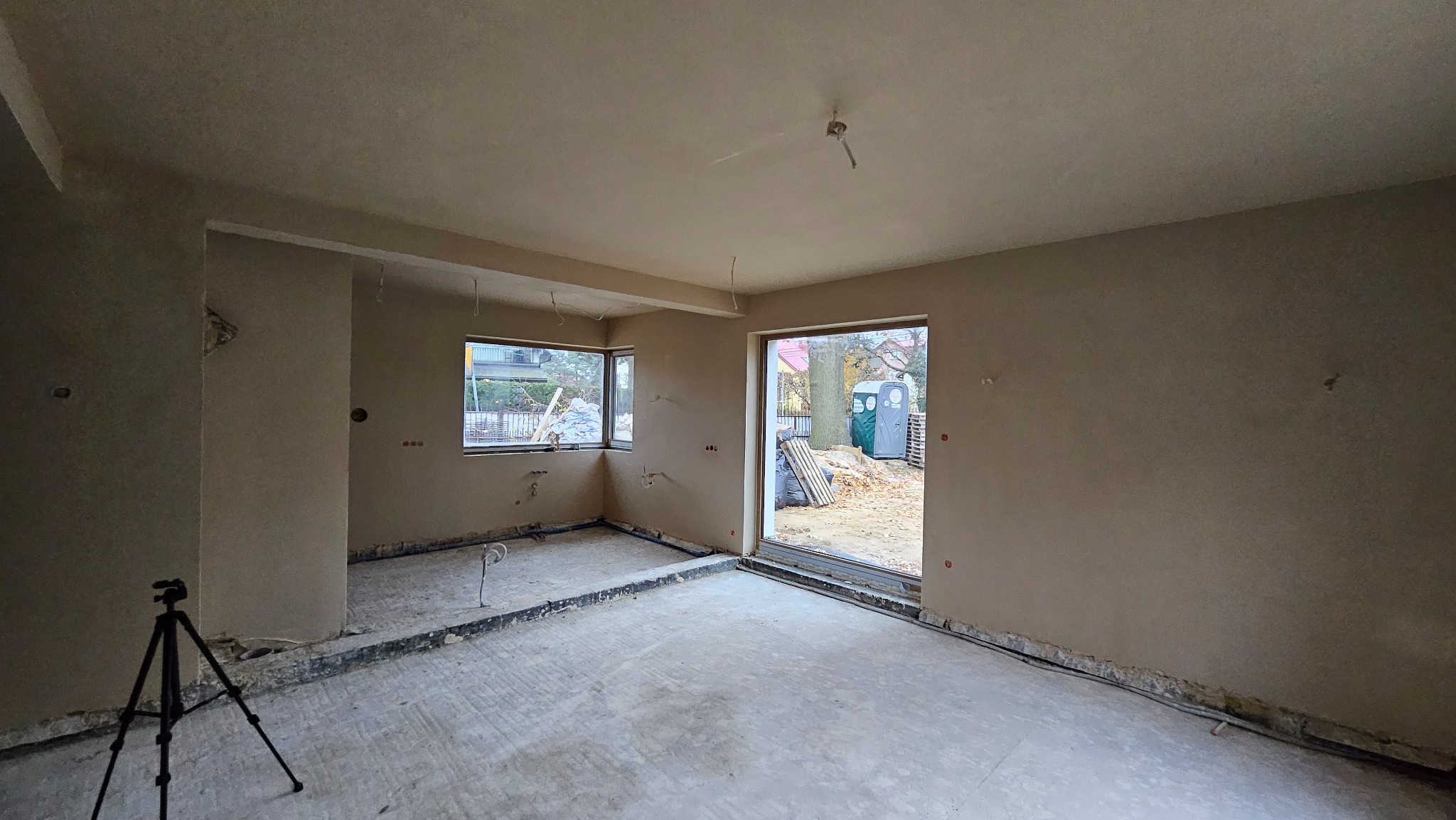 Surowe wnętrze po nałożeniu tynku, widoczne instalacje elektryczne, okno z widokiem na podwórko, na pierwszym planie statyw aparatu.