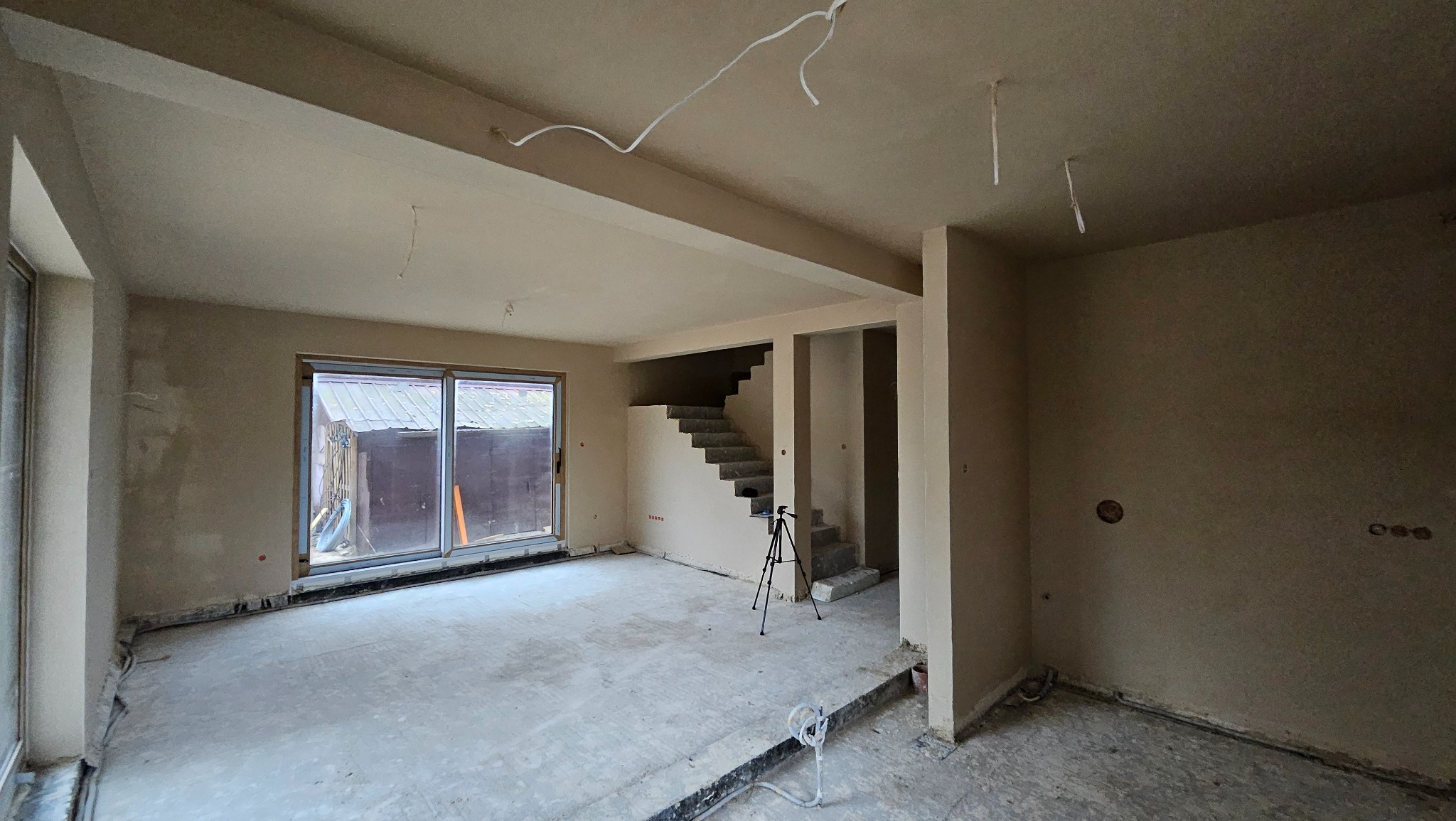 Wnętrze domu w trakcie prac wykończeniowych: ściany pokryte świeżym tynkiem, widoczne przewody elektryczne, surowa posadzka, schody w stanie surowym oraz statyw fotograficzny.