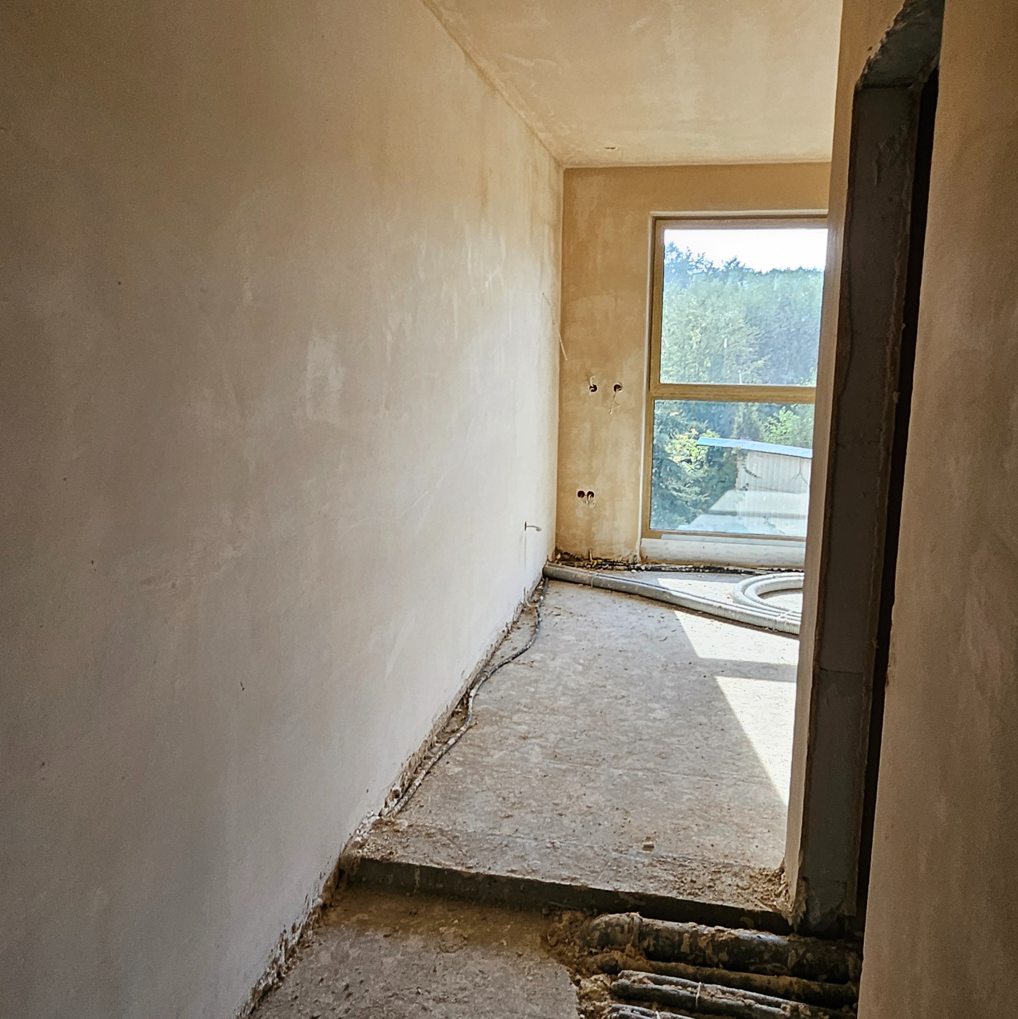 Wnętrze w trakcie prac tynkarskich: ściany pokryte świeżym tynkiem, widoczne puszki elektryczne i rury wentylacyjne przy podłodze, okno z widokiem na zieleń.