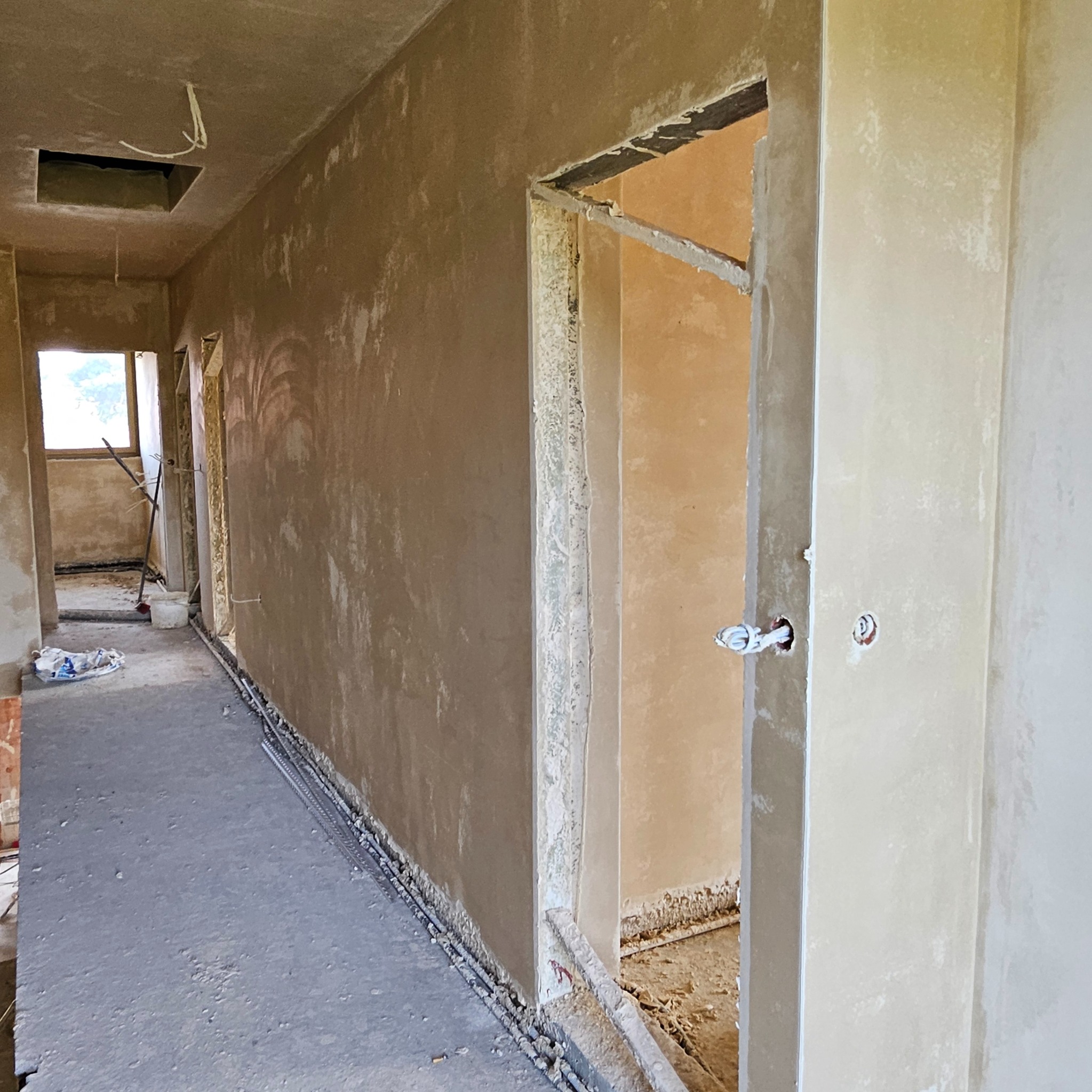 Korytarz w stanie surowym po tynkowaniu maszynowym, widoczne ślady po tynkowaniu, wystające kable, otwory w ścianach na instalacje elektryczne, podłoga betonowa ze zbrojeniami przy ścianach.