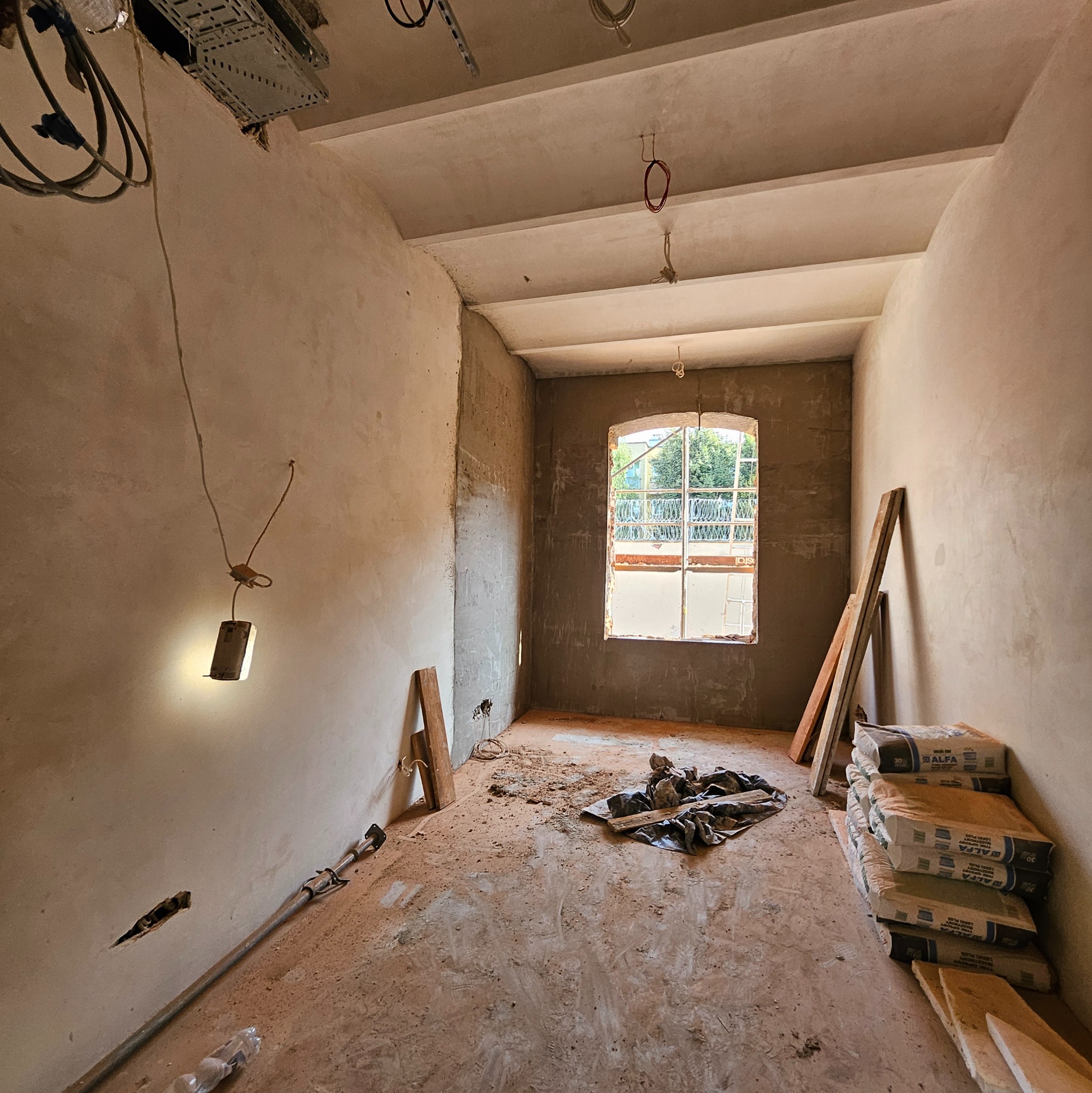 Wnętrze w trakcie prac wykończeniowych: surowe tynki na ścianach, worki z zaprawą budowlaną przy ścianie, prowizoryczne oświetlenie i okno z widokiem na zieleń.