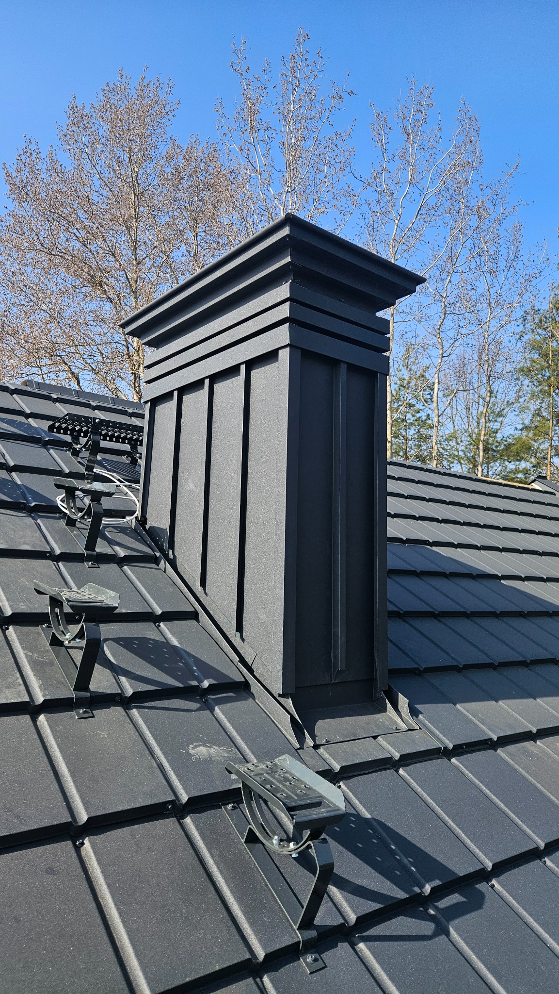 Szary dach z blachy trapezowej z zamontowanymi czarnymi stopniami kominiarskimi i kominem obłożonym blachą w słoneczny dzień.