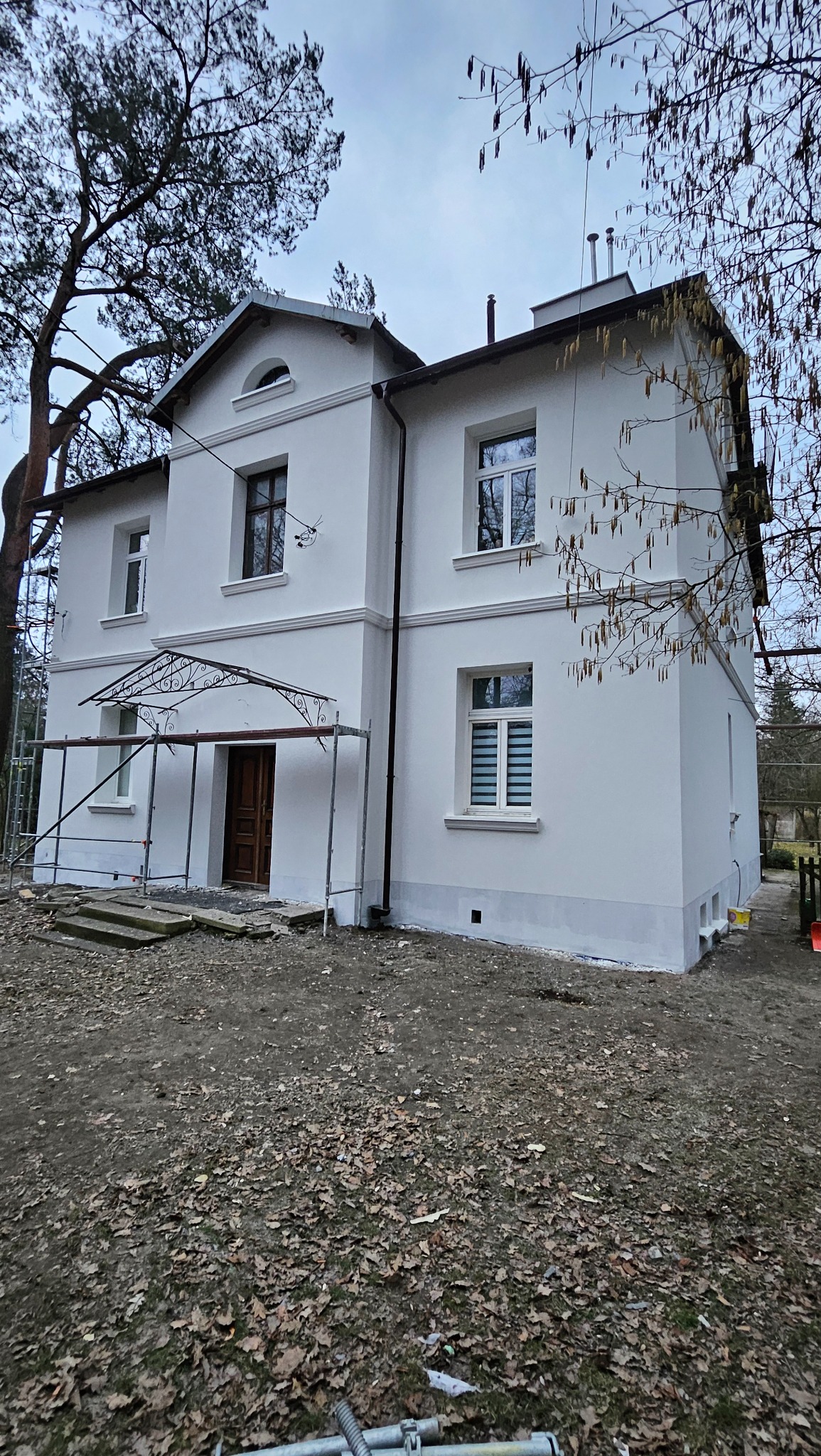 Dwupiętrowy dom z białymi ścianami po renowacji, z widocznymi śladami świeżego malowania u podstawy, z metalowym zadaszeniem nad wejściem, otoczony drzewami i rusztowaniem.