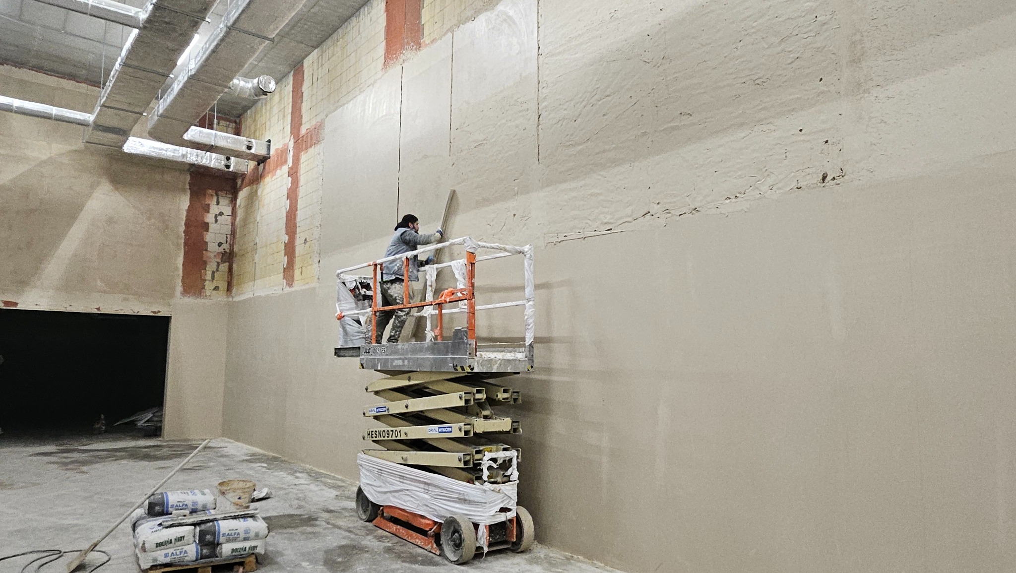 Pracownik na podnośniku nożycowym wygładza świeżo nałożony tynk na ścianie w surowym wnętrzu budynku, widoczne worki z zaprawą tynkarską.