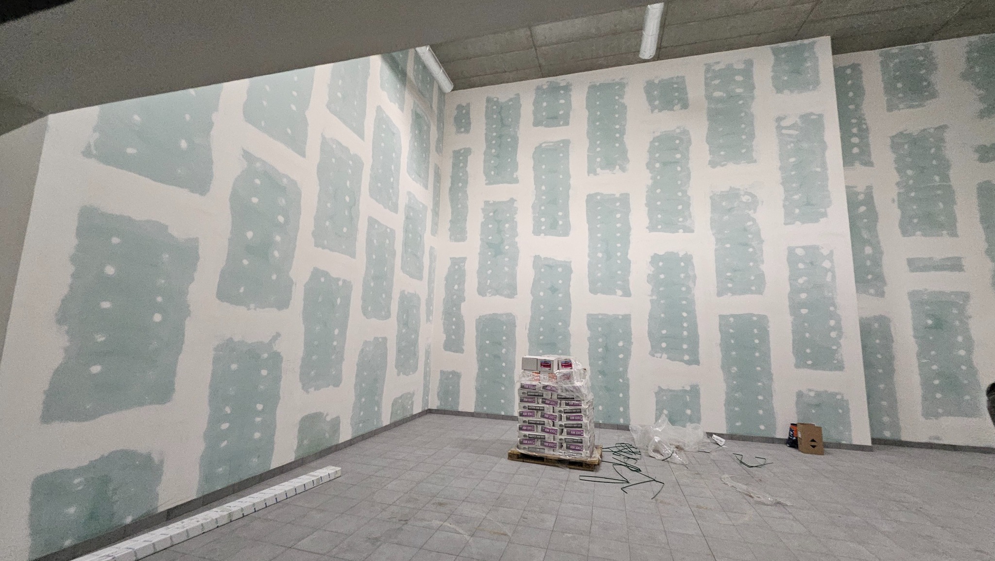 Wnętrze w trakcie tynkowania maszynowego, widoczne ściany z nałożonym tynkiem w pasach, paleta z workami tynku oraz elementy montażowe na podłodze.