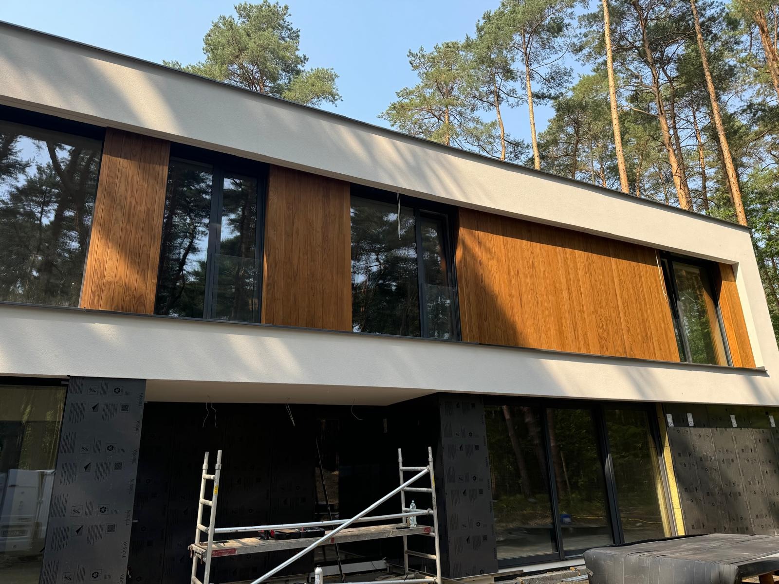 Nowoczesny dom z częściowo ocieploną elewacją, widoczne panele imitujące drewno na górnej kondygnacji oraz rusztowanie na dole, otoczenie leśne.