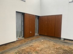 Nowoczesna elewacja budynku z brązowymi panelami drewnopodobnymi, częściowo zasłaniającymi okno, widoczne świeże ślady prac ziemnych.