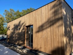 Nowoczesny dom jednorodzinny z elewacją z naturalnego drewna, widoczne okno i fragment fundamentu, jasne oświetlenie, drzewa w tle.