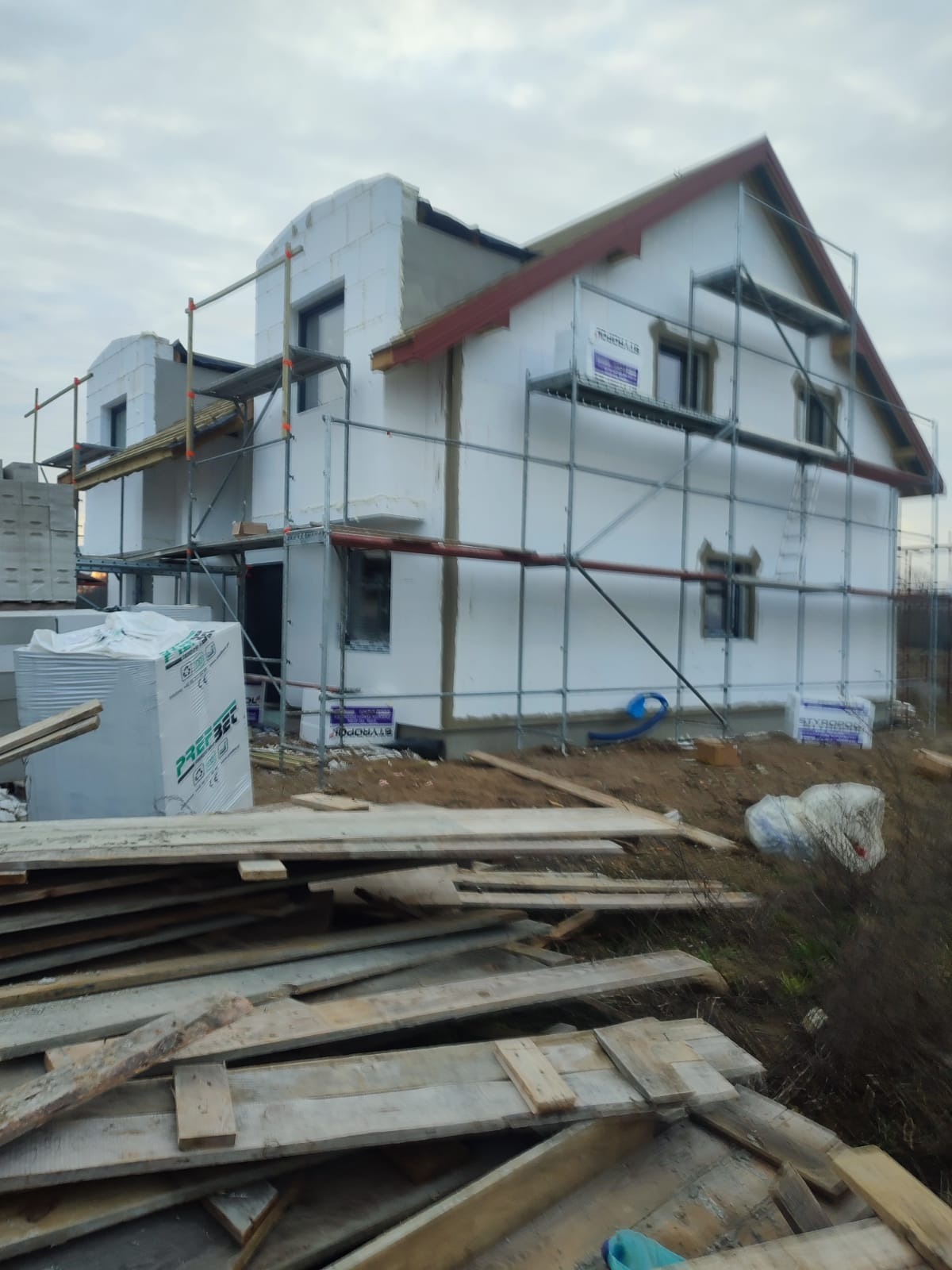 Dom w trakcie ocieplania styropianem, widoczne rusztowania, materiały budowlane i częściowo wykończony dach z czerwoną podbitką.