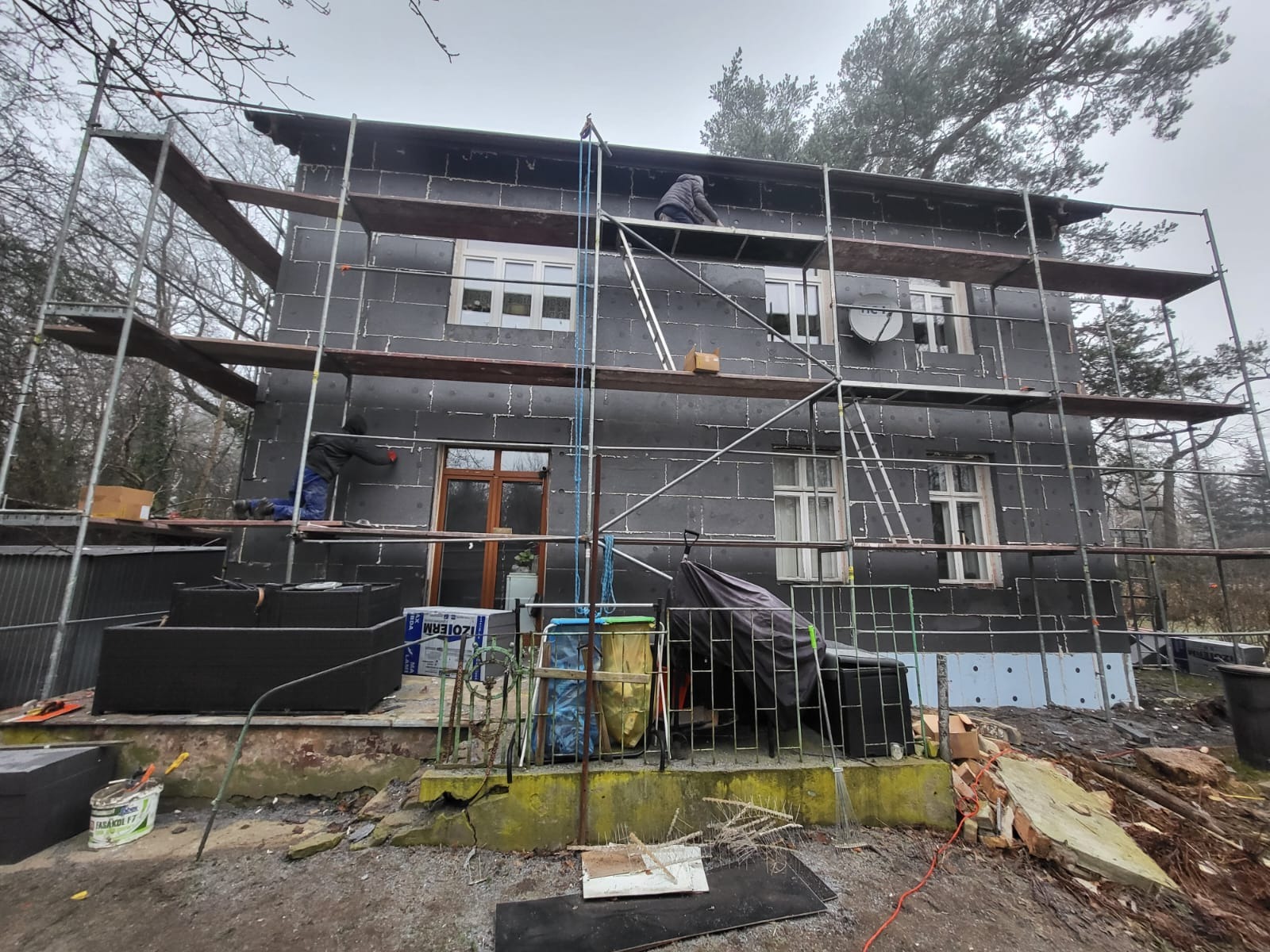 Dwupiętrowy dom w trakcie prac ociepleniowych elewacji, pokryty czarnym styropianem, z widocznymi rusztowaniami i pracownikami na rusztowaniach, podwórko z gruzem i narzędziami.