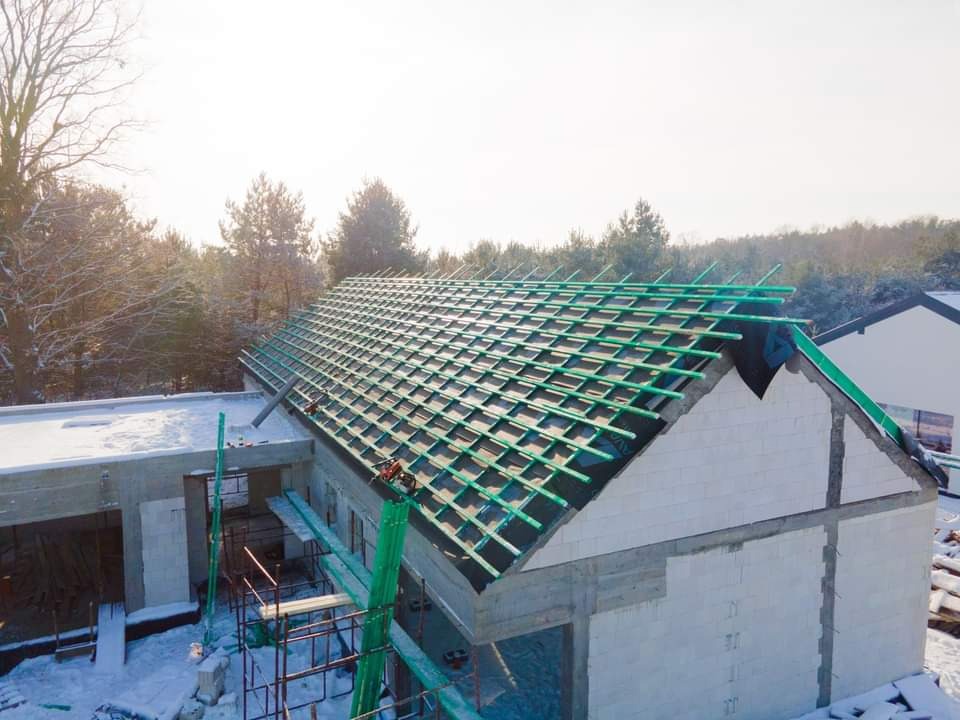 Budowa domu w zimowej scenerii: widok na nieotynkowane ściany z betonu komórkowego i konstrukcję dachu z zielonymi łatami, przygotowaną pod pokrycie.