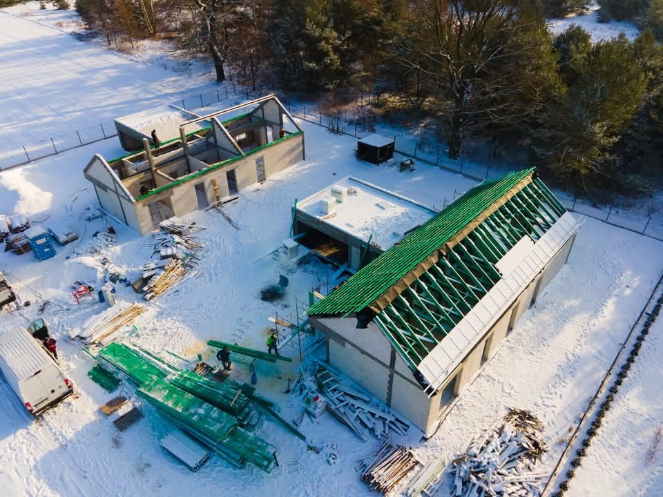 Zimowy widok z drona na plac budowy z dwoma budynkami w stanie surowym: jeden z konstrukcją dachu w trakcie montażu zielonego pokrycia, drugi w trakcie wznoszenia ścian. Śnieg pokrywa teren...
