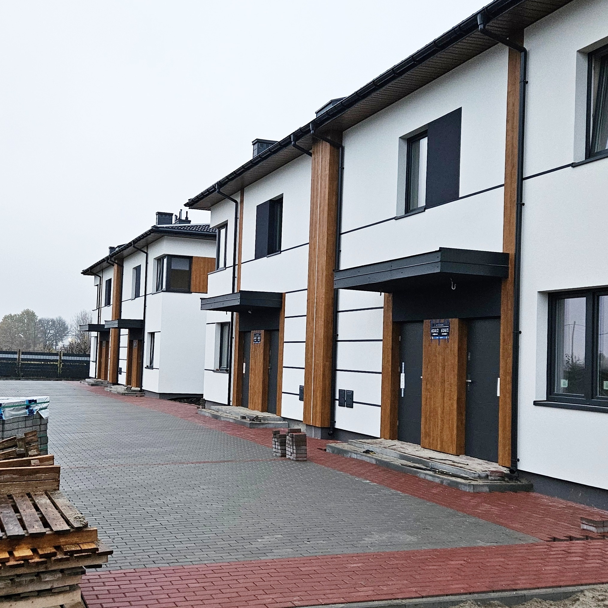 Nowoczesne segmenty bliźniacze z białą elewacją, drewnianymi akcentami i szarą stolarką okienną, widok z zewnątrz na kostkę brukową i palety z materiałami budowlanymi w tle.