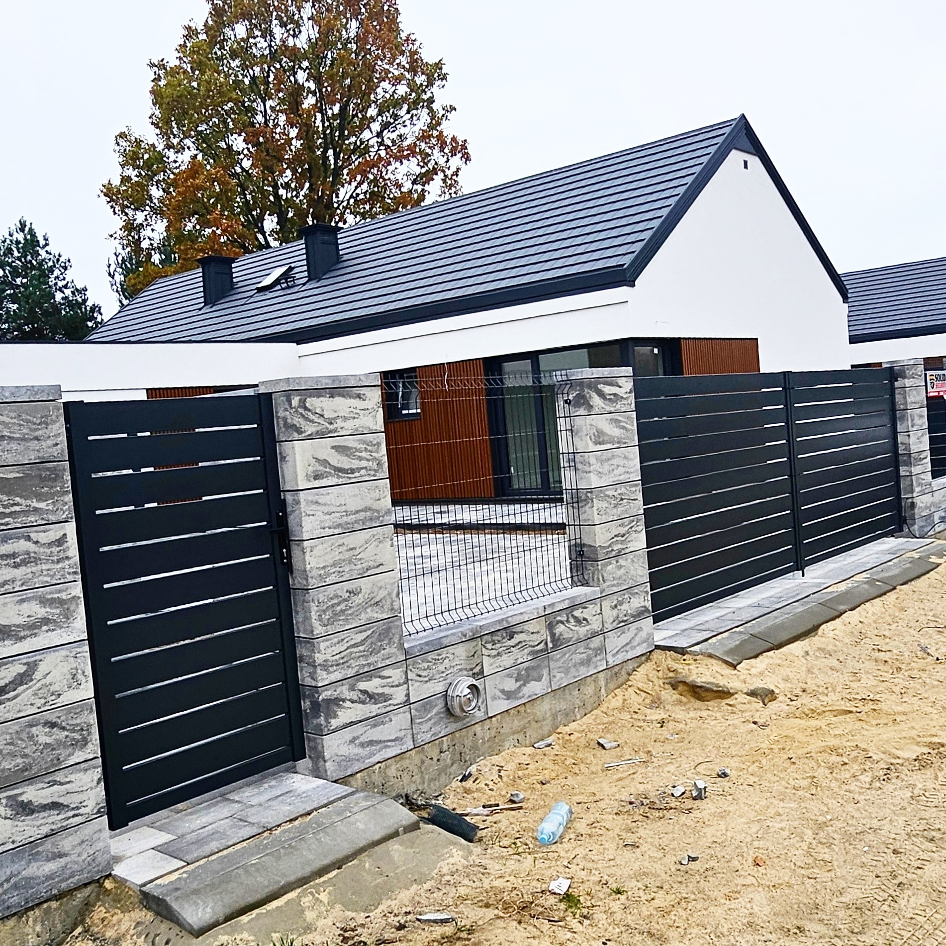 Nowoczesne ogrodzenie posesji z szarych, betonowych bloczków imitujących kamień i czarnych, poziomych paneli, widok z zewnątrz na tle domu z ciemną dachówką.