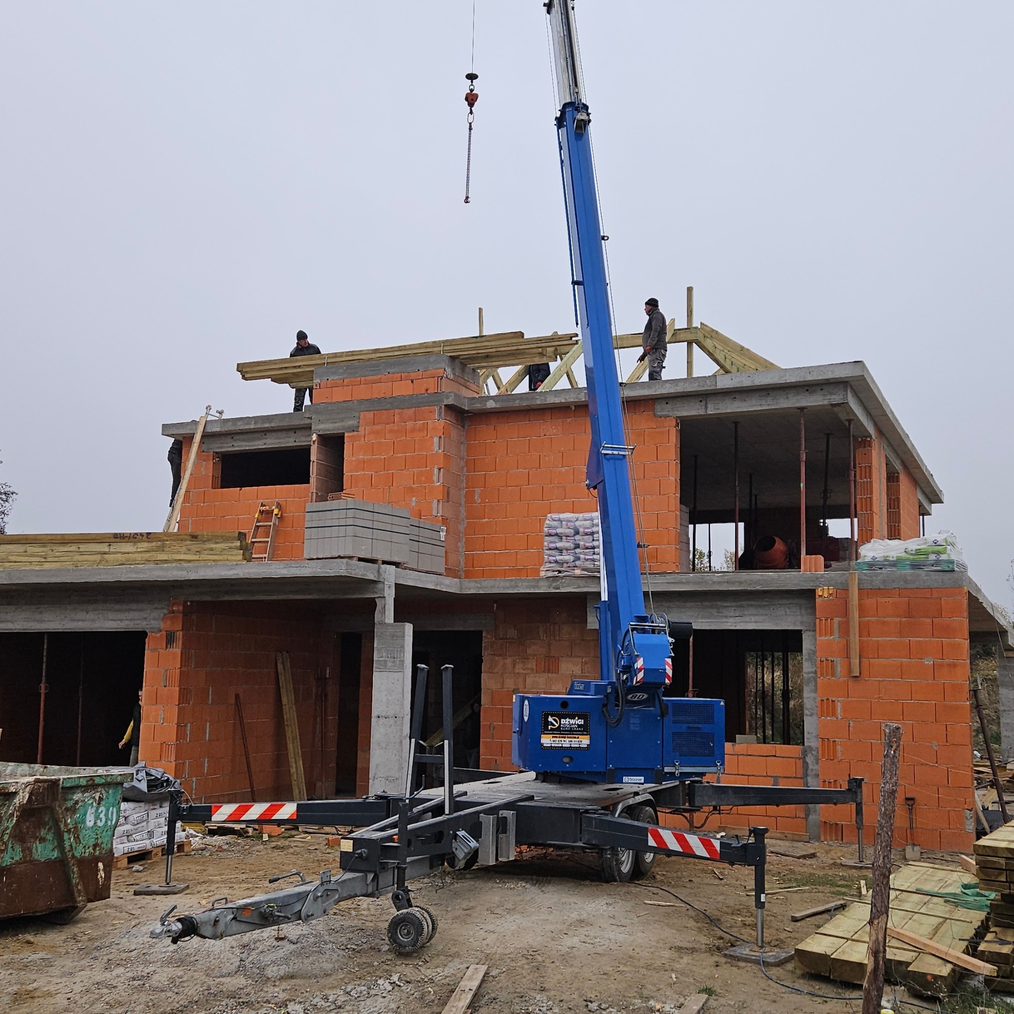 Budowa domu jednorodzinnego w stanie surowym otwartym, z widocznym dźwigiem teleskopowym, pustakami ceramicznymi i drewnianą konstrukcją dachu w trakcie montażu.