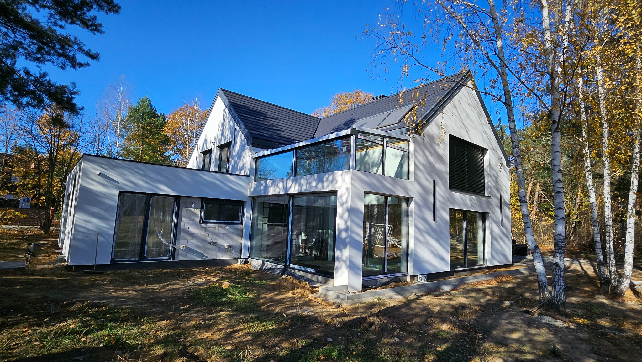 Nowoczesny dom jednorodzinny w Serocku z jasną elewacją i przeszkloną werandą, widok z zewnątrz w jesiennej scenerii.