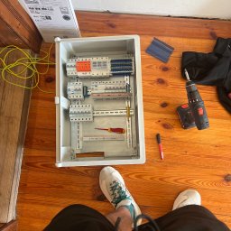 Voltex Anton Obukhov - Otwarta skrzynka rozdzielcza z zamontowanymi elementami, widok z góry na drewnianej podłodze, obok wiertarka akumulatorowa, śrubokręt i pisak, w tle fragment kartonu i para butów osoby fotografującej.