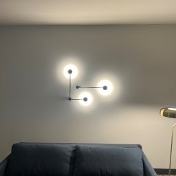 Voltex Anton Obukhov - Nowoczesna lampa ścienna z trzema kulistymi kloszami, zamontowana nad granatową sofą w pomieszczeniu o szarych ścianach, z widocznym fragmentem złotej lampy podłogowej.