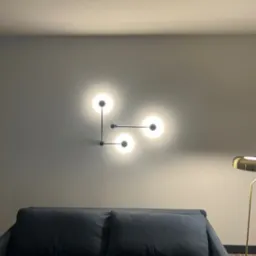 Nowoczesna lampa ścienna z trzema kulistymi kloszami, zamontowana nad granatową sofą w pomieszczeniu o szarych ścianach, z widocznym fragmentem złotej lampy podłogowej.