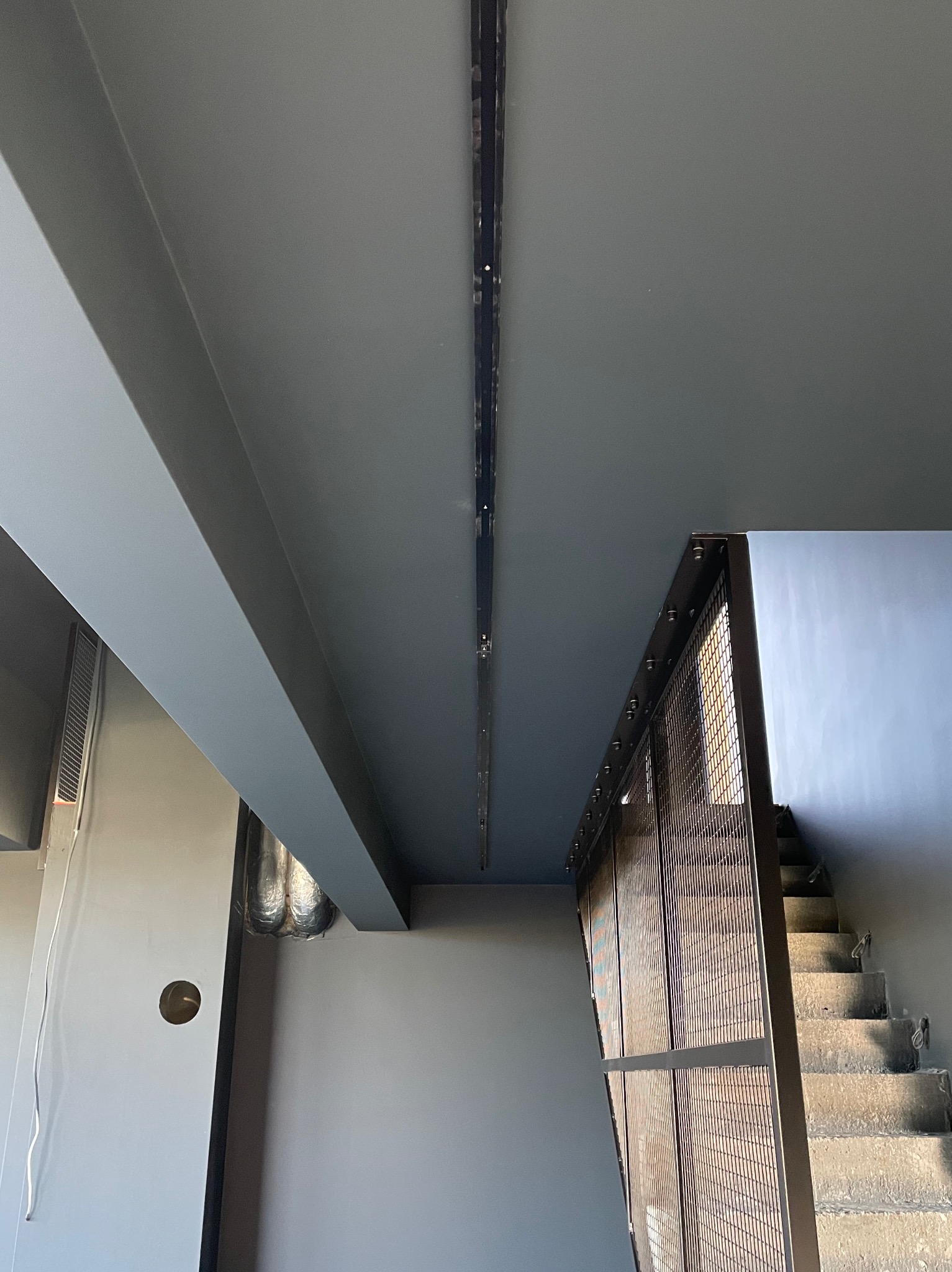 Szara klatka schodowa z betonowymi schodami, metalową balustradą z siatki i liniowym systemem oświetleniowym LED zamontowanym na suficie.