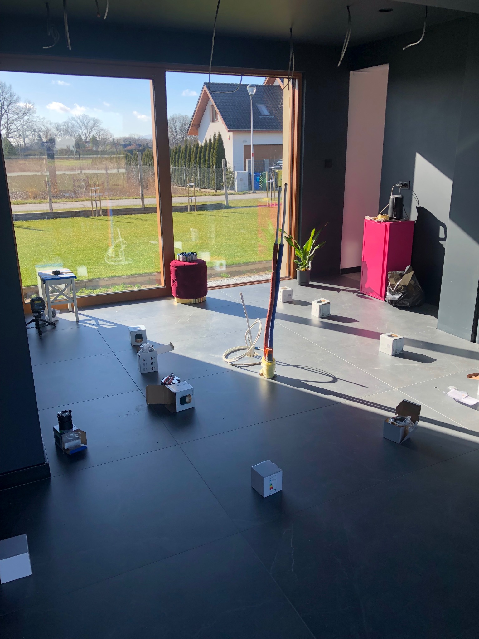 Wnętrze w trakcie montażu oświetlenia: widoczne kable wystające z sufitu, pudełka po lampach rozmieszczone na podłodze, okno z widokiem na ogród.