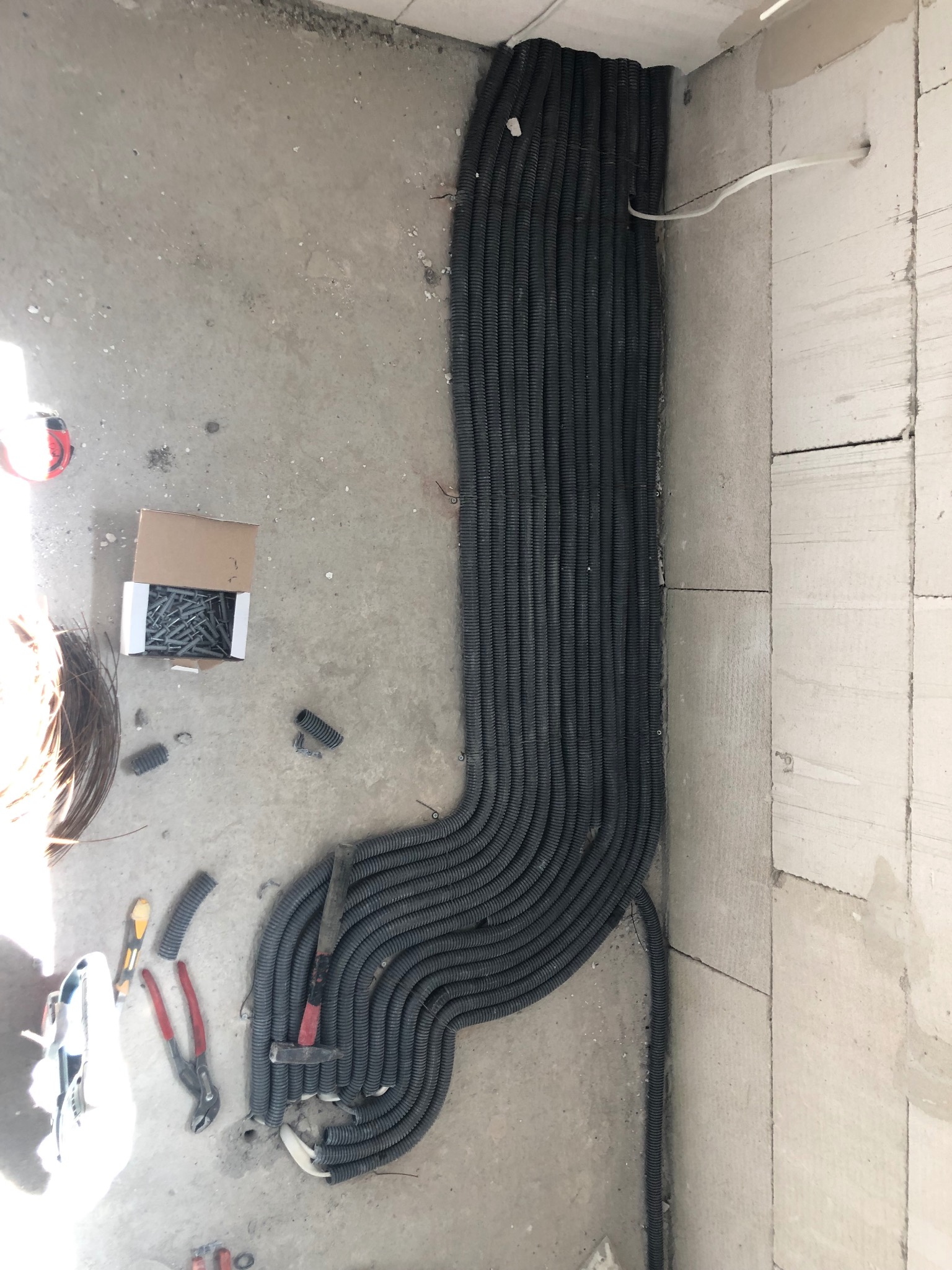 Rury karbowane ułożone wzdłuż surowej ściany, narzędzia i pudełko z wkrętami na podłodze w trakcie instalacji.