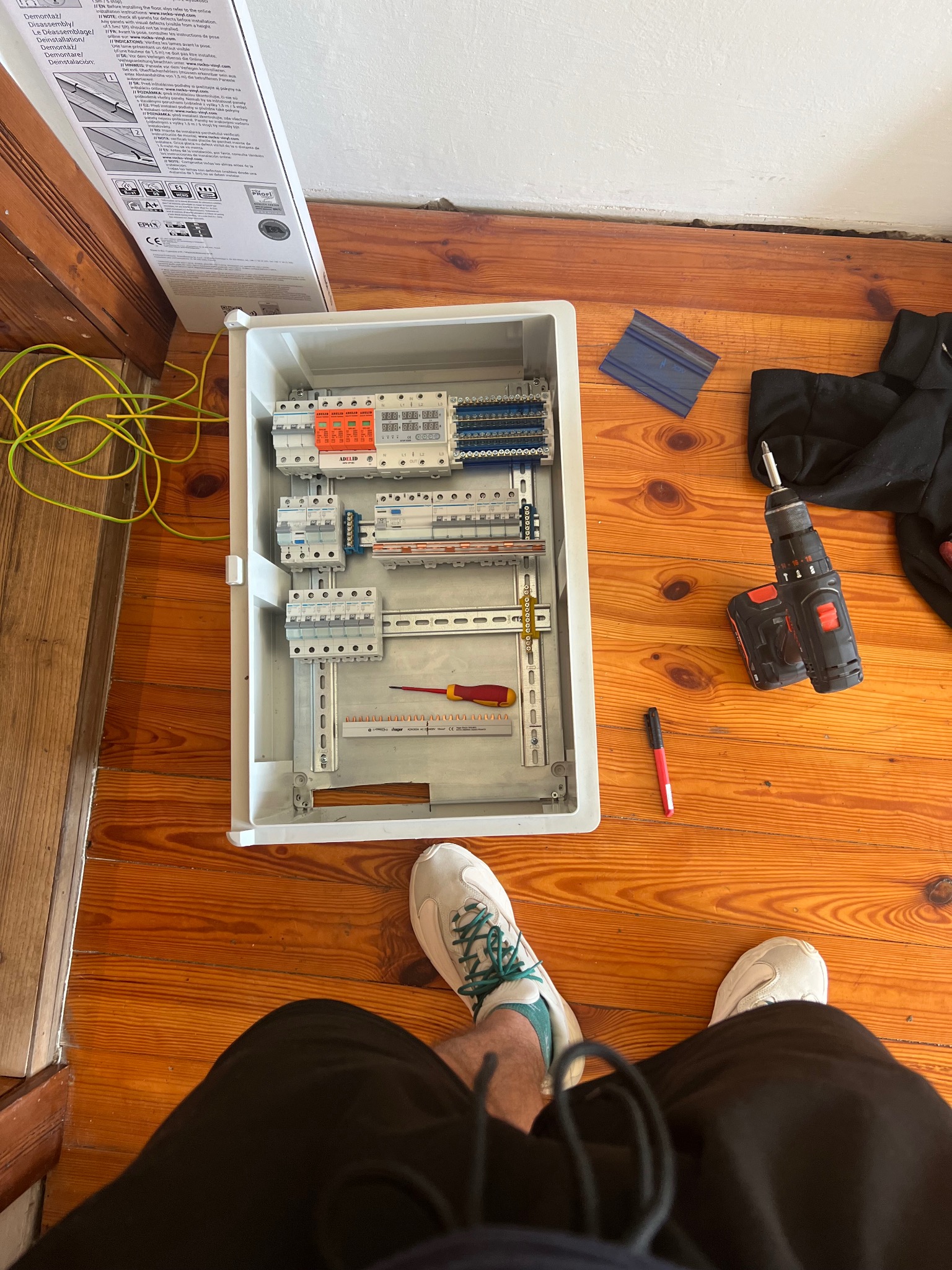 Otwarta skrzynka rozdzielcza z zamontowanymi elementami, widok z góry na drewnianej podłodze, obok wiertarka akumulatorowa, śrubokręt i pisak, w tle fragment kartonu i para butów osoby fotografującej.