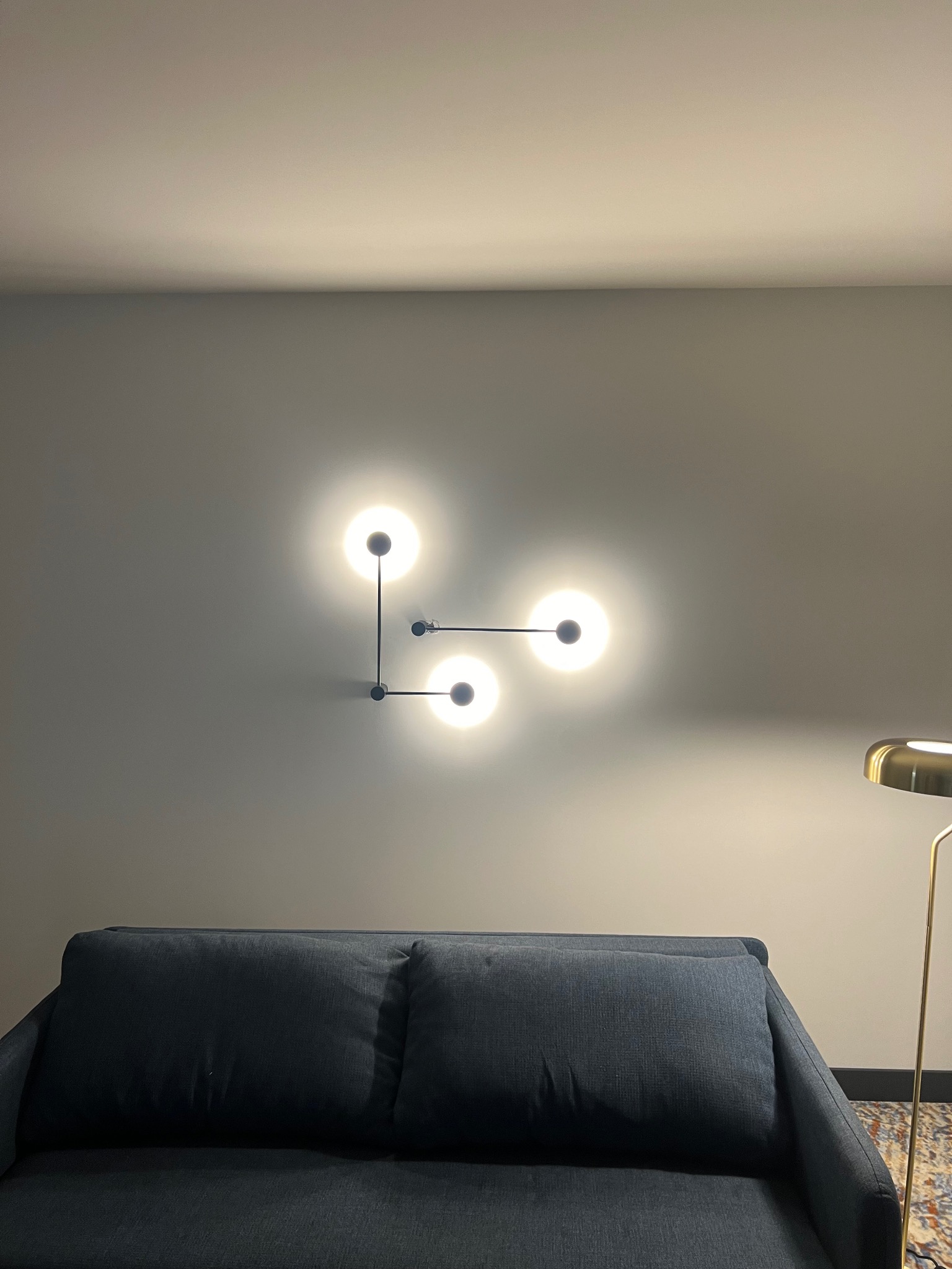 Nowoczesna lampa ścienna z trzema kulistymi kloszami, zamontowana nad granatową sofą w pomieszczeniu o szarych ścianach, z widocznym fragmentem złotej lampy podłogowej.
