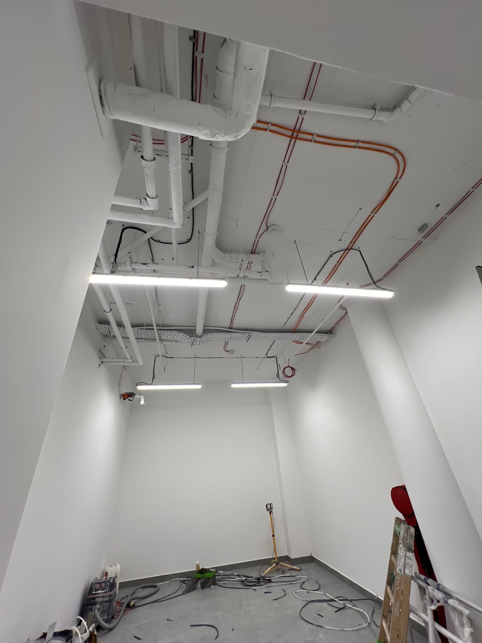 Wnętrze w trakcie remontu z widocznymi instalacjami pod sufitem: rurami, kablami w kolorach pomarańczowym i czerwonym, oraz zamontowanymi lampami liniowymi. Na podłodze leżą kable i narzędzia, a...