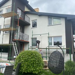 Usługi remontowe-budowlane - Malowanie elewacji domu na rusztowaniu. Dwóch malarzy przy pracy, widoczne pędzle, farba, zabezpieczone balkony folią. Ciemne akcenty architektoniczne.