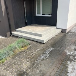 Usługi remontowe-budowlane - Nowoczesne schody wejściowe z jasnoszarej kostki brukowej, prowadzące do domu z czarną fasadą i białym fragmentem ściany, otoczone zielenią i kostką na podwórku.