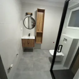 Nowoczesna łazienka z szarymi płytkami, drewnianą szafką pod umywalką, okrągłym lustrem i kabiną prysznicową z czarną ramą. Minimalistyczny design.