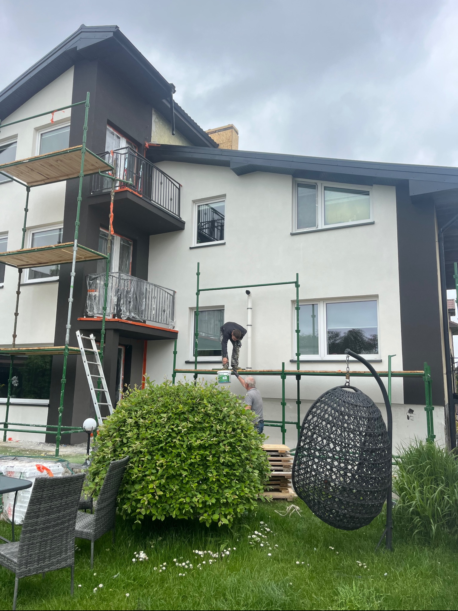 Malowanie elewacji domu na rusztowaniu. Dwóch malarzy przy pracy, widoczne pędzle, farba, zabezpieczone balkony folią. Ciemne akcenty architektoniczne.