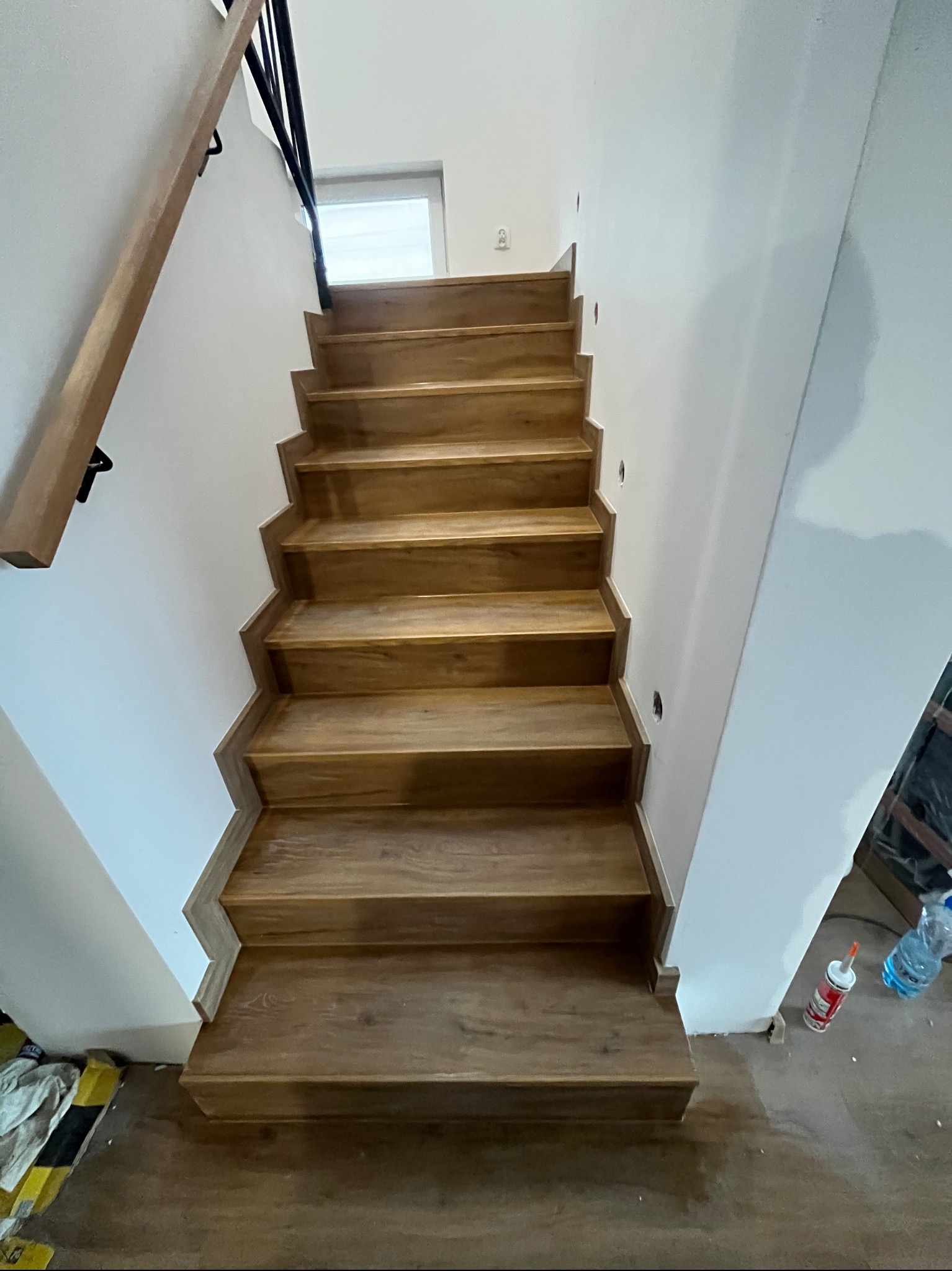 Drewniane schody z czarną balustradą w jasnym wnętrzu. Widoczne wykończenia i detale, perspektywa od dołu. Elementy montażowe na podłodze.