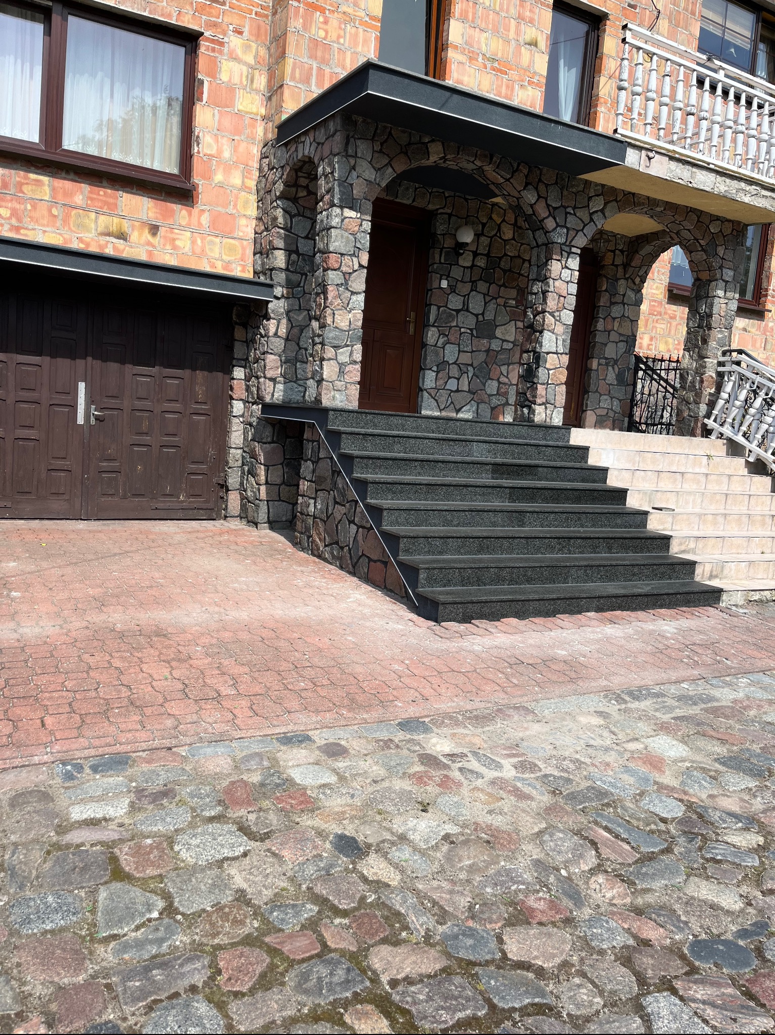 Wykończenie schodów wejściowych z ciemnego kamienia w Minkowicach. Kamienne obłożenie budynku i podjazdu z kostki. Estetyczne detale architektoniczne.