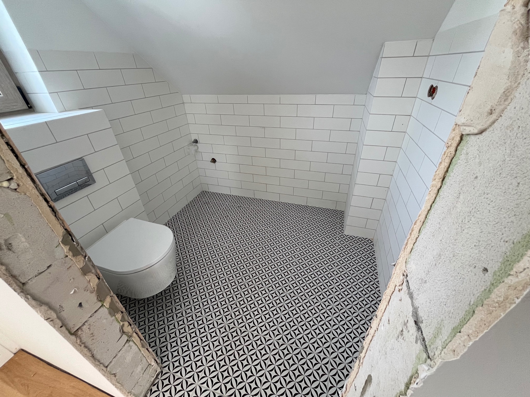 Biała toaleta i wzorzysta podłoga w łazience na poddaszu. Ściany wyłożone białymi, prostokątnymi płytkami. Fragment ściany w surowym stanie.