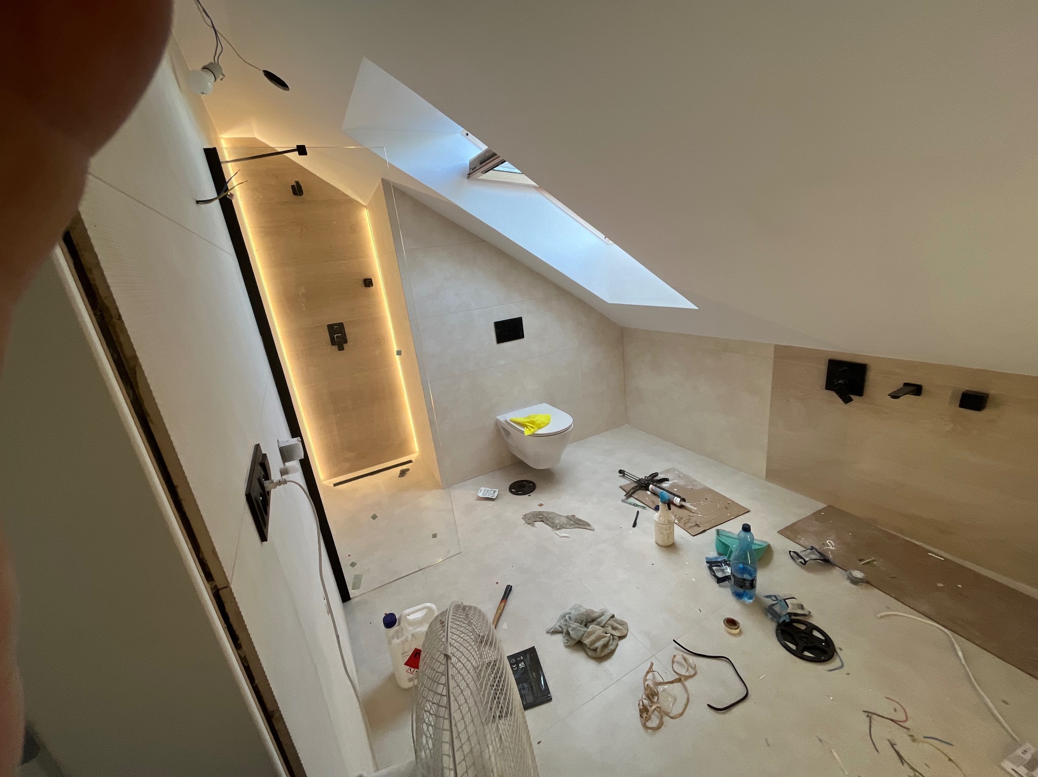 Remontowana łazienka na poddaszu ze skośnym oknem dachowym. Kabina prysznicowa z podświetleniem LED, biała toaleta i liczne narzędzia na podłodze.