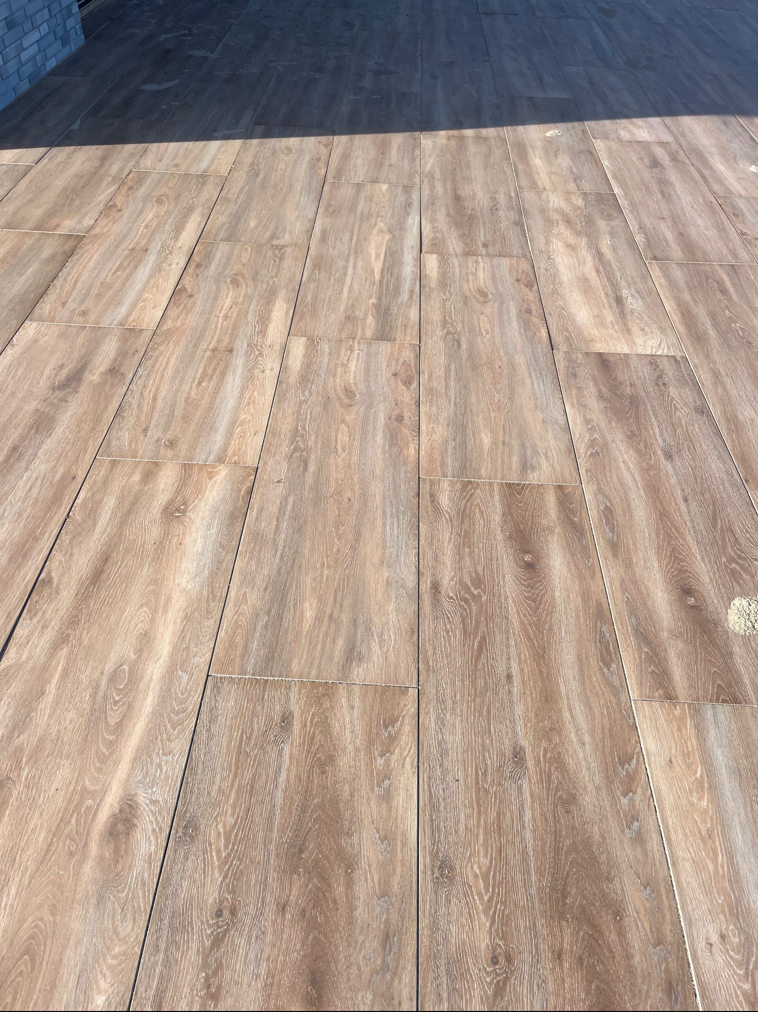 Ułożona podłoga z płytek imitujących drewno, w ciepłych odcieniach brązu. Widoczne regularne spoiny i fragment ściany w tle, w górnej części kadru cień.