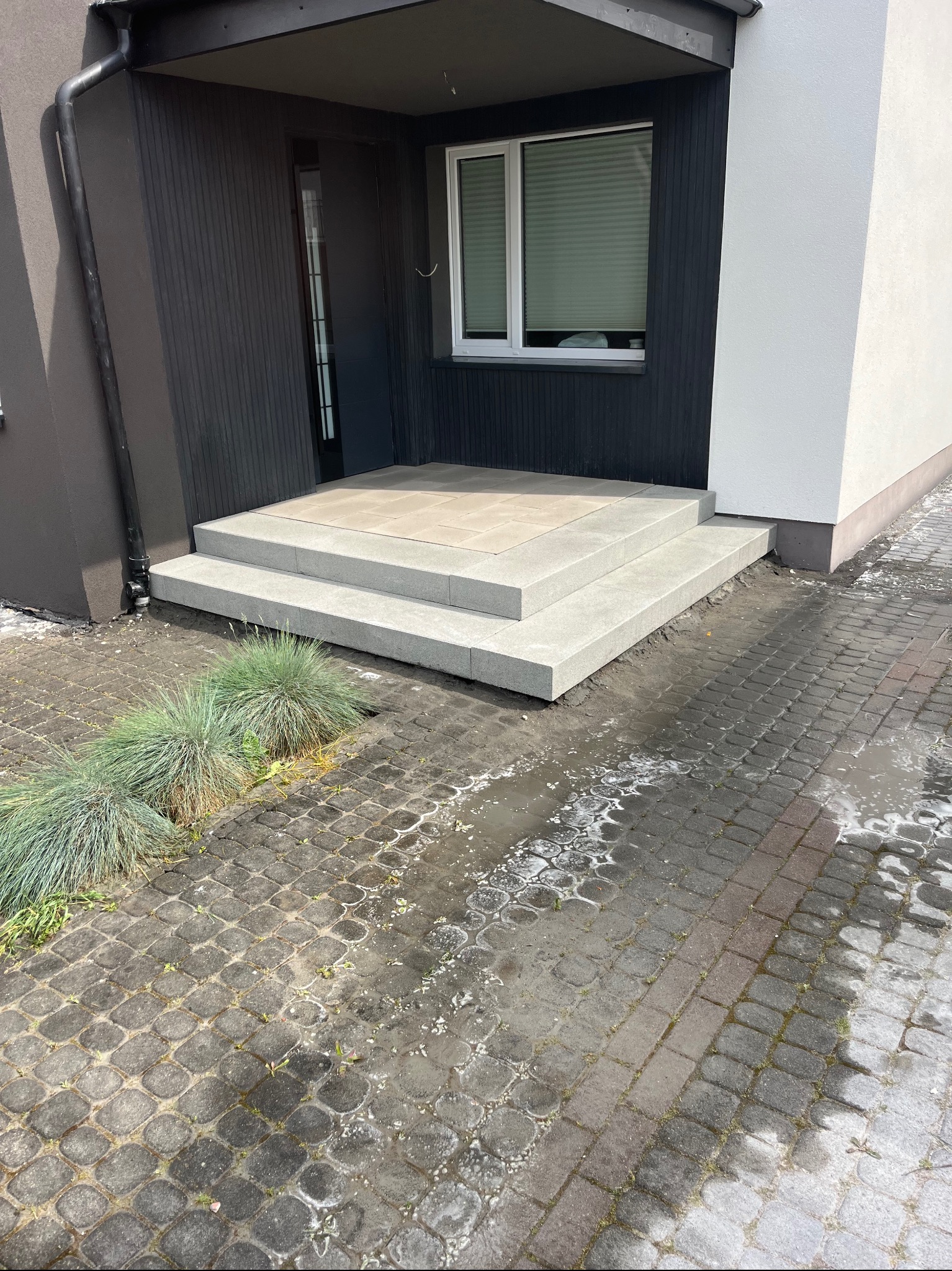 Nowoczesne schody wejściowe z jasnoszarej kostki brukowej, prowadzące do domu z czarną fasadą i białym fragmentem ściany, otoczone zielenią i kostką na podwórku.