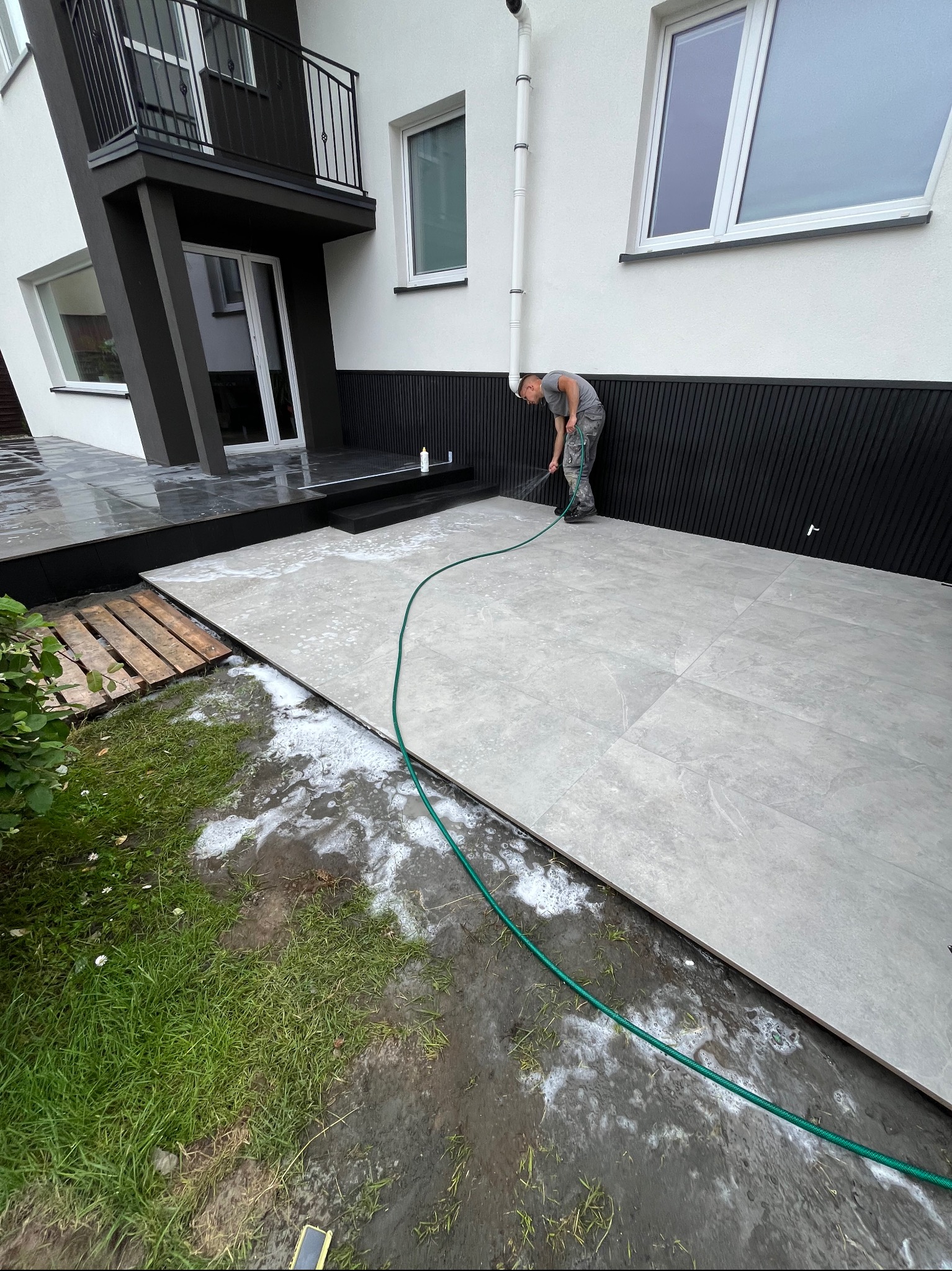 Mycie ciśnieniowe nowej, szarej, dużej kostki brukowej przy nowoczesnym domu z czarną fasadą. Widoczny pracownik z wężem i pianą czyszczącą.