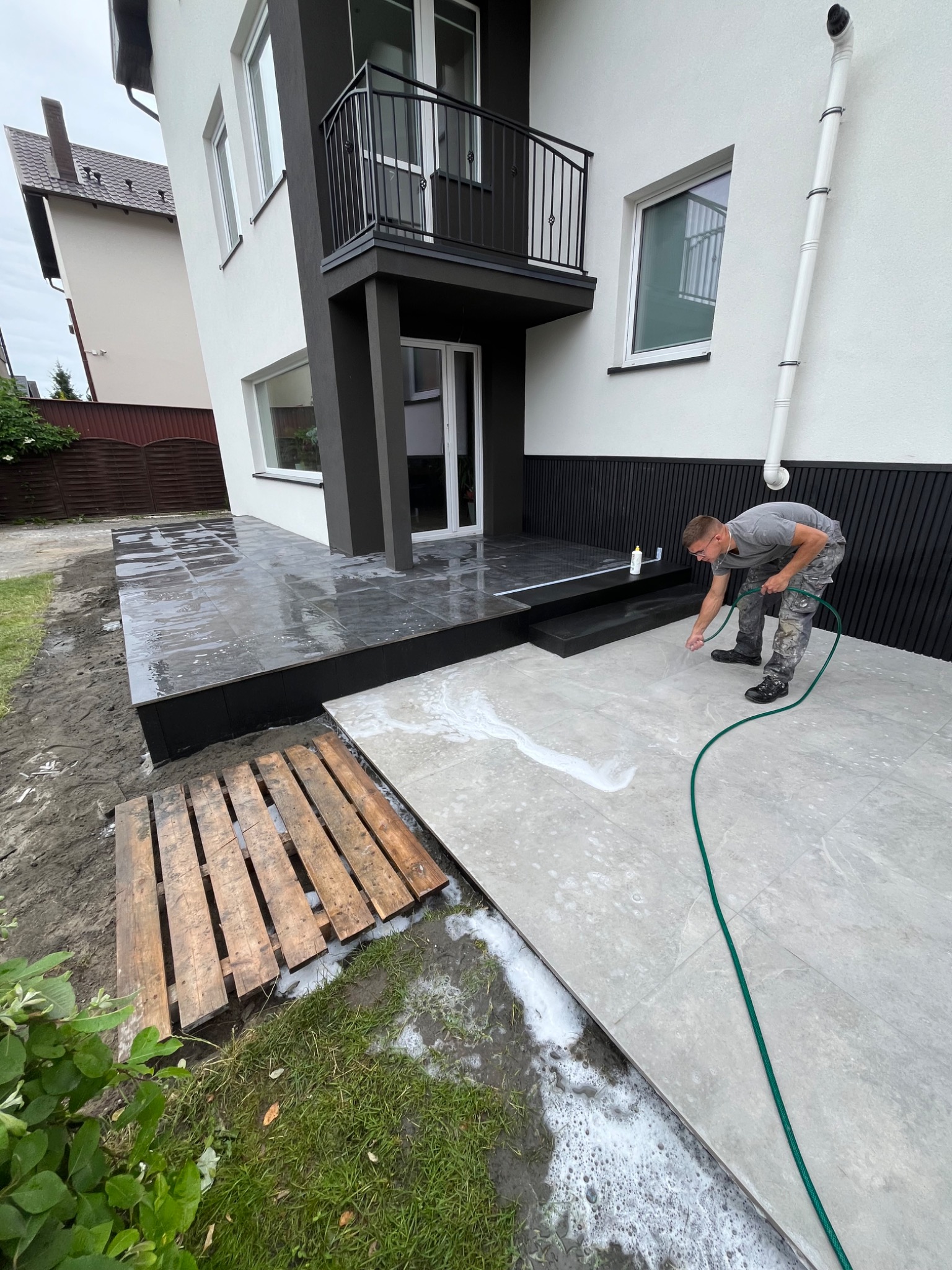 Czyszczenie płytek tarasowych szarym płynem pod ciśnieniem. Paleta drewniana obok. Nowoczesny dom z balkonem w tle.